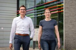 Tobias Erb und Anke Becker, Sprecher und Sprecherin des Exzellenzclusters M4C gehen in Freizeitkleidung nebeneinander vor dem SYNMIKRO Gebäude entlang. Im Hintergrund bunte Streifen über dem Gebäudeeingang.