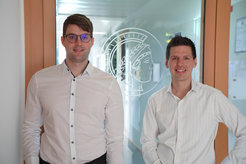 Two men in white shirts are standing in front of a glass door. On the glass door is an emblem with an ancient head and laurel wreath, Minerva, the logo of the Max Planck Society.